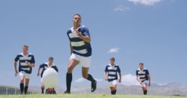 Ragbi topunu tekmeleyen karışık ırktan bir erkek rugby oyuncusunun düşük açılı görüntüsü, arkasında bir grup beyaz erkek takım arkadaşı duruyor, hepsi de takım elbisesi giyiyor, bir ragbi sahasında, ağır çekimde, ağır çekimde.