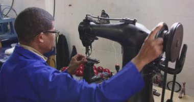 Bir fabrikada çalışan, kriket topları yapan, deri kaplamaları diken ve iplikleri el aletleriyle tamamlayan endüstriyel dikiş makinesi kullanan Afrika kökenli Amerikalı bir adamın yan görüntüsü.