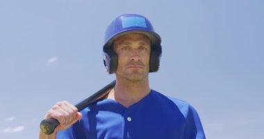 Kafkasyalı bir erkek beyzbol oyuncusu, bir vurucu, takım üniforması ve kaskı giyiyor, spor sahasında antrenman yapıyor, yavaş çekimde beyzbol sopası tutuyor.