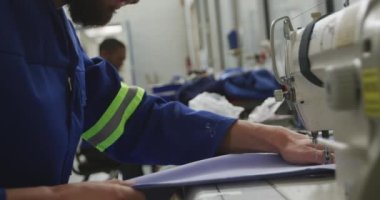 Bir fabrikadaki bir atölyede sakallı, tekerlekli sandalye yapan, bir tezgahta oturan, dikiş makinesi kullanan, başka bir işçiyle birlikte ağır çekimde batakhanede dikiş makinesi kullanan beyaz bir erkek işçinin yan görüntüsü.