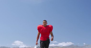 Melez bir Amerikan futbolcusunun kaskını tutarken ve spor sahasında ağır çekimde kameraya doğru yürürken alçak açılı görüntüsü. Stadyumda Koşu ve Spor Eğitimi.