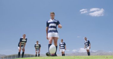 Kafkasyalı bir erkek rugby oyuncusunun ragbi topuna vuruşunun düşük açılı görüntüsü, arkasında bir grup Kafkas ve karışık ırk erkek takım arkadaşı duruyor, hepsi de takım elbisesi giyiyor, bir maç sırasında, ağır çekimde, bir ragbi sahasında.