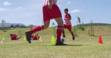 Bir grup beyaz erkek futbolcunun, takım elbiseli, güneşin altında spor sahasında antrenman yapan, külahlar arasında top süren, düşük açılı görüntüsü. Stadyumda Koşu ve Spor Eğitimi.