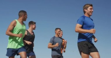Spor sahasında antrenman yapan çok ırklı bir erkek koşucu grubunun yan görüntüsü. Çim pistinde birlikte ağır çekimde koşuyorlar.