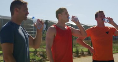 Üç beyaz erkek koşucunun spor sahasında antrenman yaparken, mola verirken, su içerken yan görüntüsü. Stadyumda ağır çekimde Koşu ve Spor Eğitimi