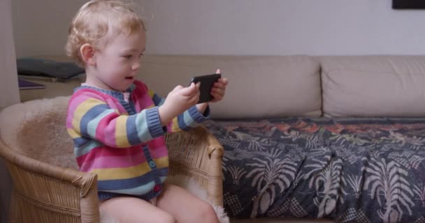 一个白人幼儿在家里享受时光 坐在客厅的椅子上 拿着和看智能手机 慢动作的侧视图 — 图库视频影像