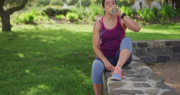 一名身穿运动服的白人年轻女子坐在矮墙上 在公园锻炼时喝水 动作缓慢 — 图库视频影像