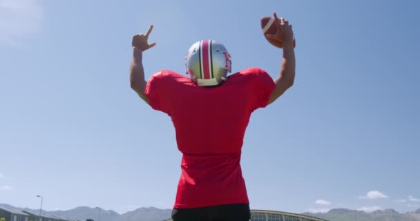 一个混血的美国足球运动员举着球 举起胳膊 在蓝天的映衬下 慢动作地指向运动场的后视镜 体育场的田径训练 — 图库视频影像