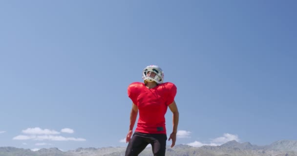 Frontansicht eines gemischten American-Football-Spielers, der in Zeitlupe vor blauem Himmel auf einem Sportplatz aufspringt und einen Ball fängt. Leichtathletik-Training im Stadion.