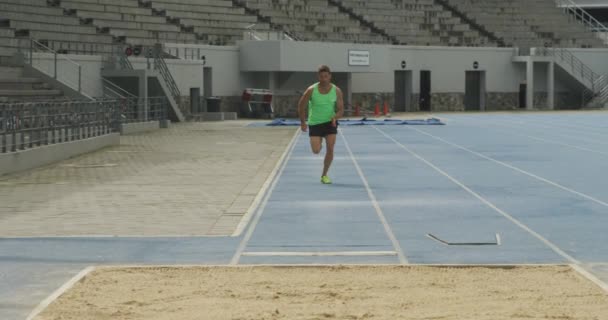 スポーツ競技場で練習している白人男性選手のフロントビュー 長いジャンプを行います スローモーション 陸上競技場での陸上競技トレーニング — ストック動画