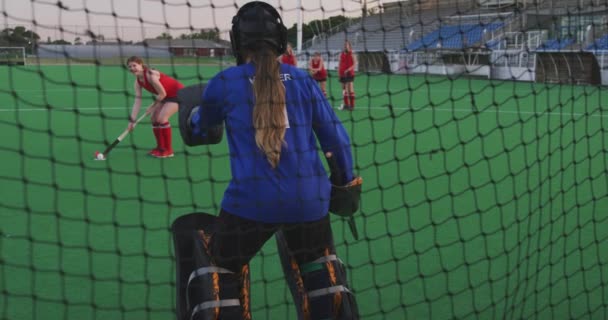 10代の白人女性ホッケーチームのゴールキーパーの背面ビューがピッチ上で動作し スポーツスタジアムでのホッケー試合中のゴールを守り スローモーション — ストック動画