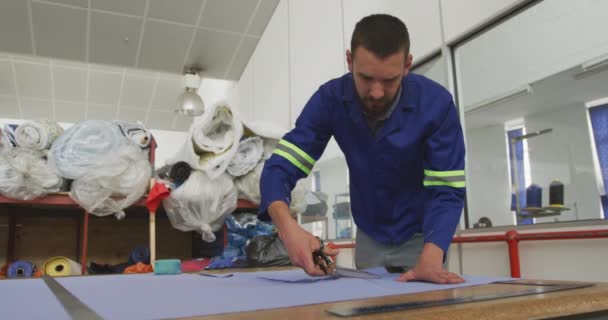 一个白人男工站在一家制造轮椅的工厂的车间里 站在工作台旁 用剪刀剪断材料 一个同事慢吞吞地走在他的面前 — 图库视频影像