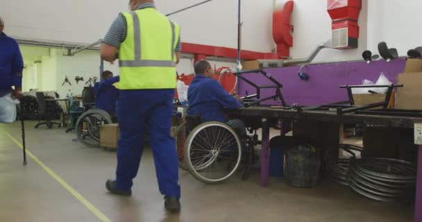 两个非洲裔美国男性工人坐在一家制造轮椅的工厂的车间里 坐在组装部分产品的工作台旁 坐在轮椅上 两个同事走过去 一个拿着拐杖 — 图库视频影像