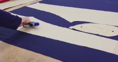 Yan görüntüde, bir spor giyim fabrikasında çalışan, mavi kumaş üzerindeki kağıt desenleri işaretleyen melez bir kadın görülüyor.