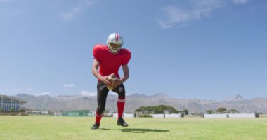 Melez bir Amerikan futbolcusunun mavi gökyüzüne karşı ağır çekimde futbol topunu tutuşu ve tekmeleyişi. Stadyumda Koşu ve Spor Eğitimi.