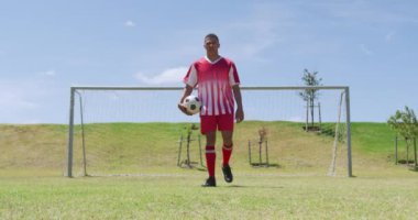 Front view of mixed-raced male football player wearing a team strip, training at a sports field in the sun, standing in front of a goal holding a football and looking to camera, in slow motion. Track and Field Sports Training in Stadium.