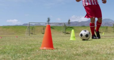 Kafkasyalı erkek futbolcunun yakın çekimde takım elbisesi giymesi, güneşte spor sahasında antrenman yapması koniler arasında ağır çekimde top sürmesi.