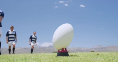 Kafkasyalı bir erkek rugby oyuncusunun, ragbi topunu tekmelerken yakın açıyla, arkasında bir grup Kafkas ve karışık ırk erkek takım arkadaşı duruyor, hepsi de ağır çekimde bir ragbi sahasında takım elbisesi giyiyor.