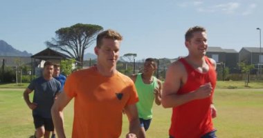 Spor sahasında antrenman yapan çok ırklı bir erkek koşucu grubunun ön görüntüsü, çimen pistinde birlikte ağır çekimde koşuyorlar.