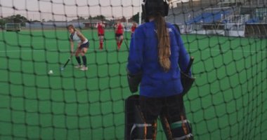 Beyazlı genç bir hokey takımının kalecisinin arka görüntüsü sahada, bir spor stadyumundaki hokey maçında kaleyi savunuyor, yavaş çekim.