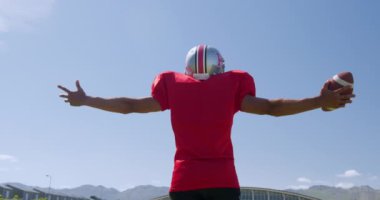 Bir spor sahasında, kollarını mavi gökyüzüne doğru açarak ağır çekimde top tutan melez bir Amerikan futbolcusunun arka görüntüsü. Stadyumda Koşu ve Spor Eğitimi.