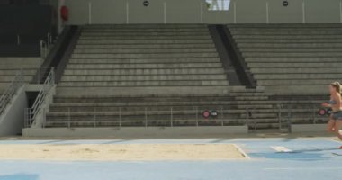 Beyazlı bayan bir sporcunun spor stadyumunda uzun atlama ve ağır çekimde antrenman yaparken yan görüntüsü. Stadyumda Koşu ve Spor Eğitimi.