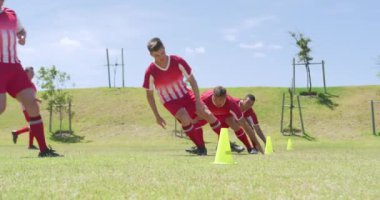 Beyaz erkek futbol oyuncularından oluşan bir takımın yan görüntüsü, takım elbiseli, güneşin altında bir spor sahasında antrenman yapan, külahlar arasında koşan, yere dokunan ve dönen, ağır çekimde. Stadyumda Koşu ve Spor Eğitimi.