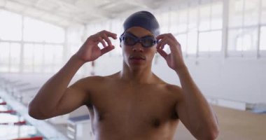 Portrait close up of mixed race male swimmer at swimming pool, wearing a blue swimming cap and swimming goggles, lifitng up his goggles and looking to camera, in slow motion