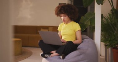 Front view of mixed race woman working in a creative office, sitting with legs crossed on a bean bag, using laptop computer, slow motion