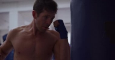 Side view close up of a Caucasian male boxer in a boxing gym punching a punchbag, slow motion. Tough Mixed Martial Arts Training.