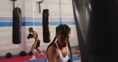 Bir boks salonunda uzun, koyu renk saç eğitimi almış, boks eldiveni giymiş ve kum torbasını yumruklamış, arka planda antrenman yapan bir kadın boksörün ön görüntüsü.