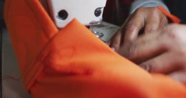 Spor giyim fabrikasında turuncu kumaş dikmek için dikiş makinesi kullanan bir kadının ellerini kapat.