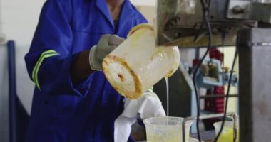 Ön planda, eldivenli, tulumlu ve şapkalı bir Afrikalı Amerikalı adam var. Fabrikada çalışıyor. Kriket topları yapıyor.