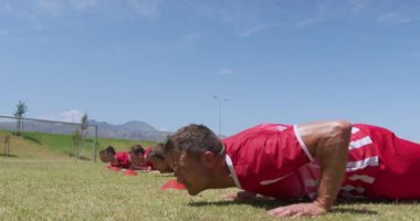 Beyaz erkek futbol oyuncularından oluşan bir takımın yan görüntüsü. Takım elbiseli, tek sıra halinde. Spor sahasında ağır çekimde şınav çekiyorlar. Stadyumda Koşu ve Spor Eğitimi.