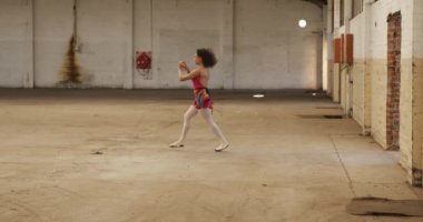 Side view of a mixed race female ballet dancer practicing in an empty warehouse, running and jumping, slow motion. Cool Generation Z hipster style concept.