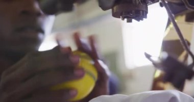 Bir fabrikada çalışan, kriket topları yapan, altın folyo detaylarını sarı bir kriket topuna aktaran bir makineyi çalıştıran Afrikalı Amerikalı 'nın yan görüntüsü.