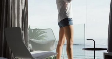 Side view of a Caucasian woman enjoying quality time in a hotel, standing on balcony drinking coffee with sea in the background. slow motion