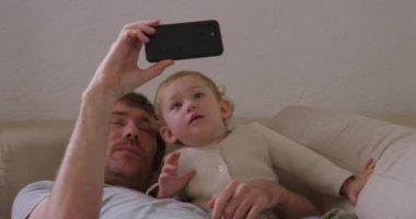 Kafkasyalı bir adamın evinde, bebeğiyle bir kanepede uzanmış, akıllı telefonuyla selfie çekerken, kucaklaşırken ve gülümserken, ağır çekimde ailece vakit geçirmesinin ön görüntüsü.