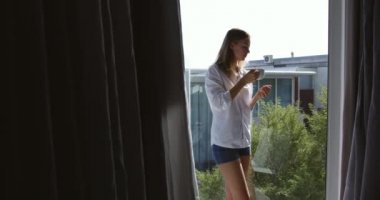Side view of a Caucasian woman enjoying quality time in a hotel, standing on balcony drinking coffee. building in the background, slow motion