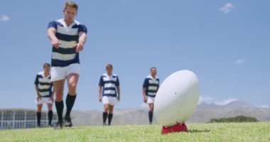 Kafkasyalı bir erkek rugby oyuncusunun ragbi topuna vuruşunun düşük açılı görüntüsü, arkasında bir grup Kafkas ve karışık ırk erkek takım arkadaşı duruyor, hepsi de takım elbisesi giyiyor, bir maç sırasında, ağır çekimde, bir ragbi sahasında.