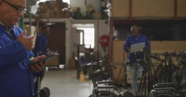 Arka planda dizüstü bilgisayar kullanan, tekerlekli sandalye üreten, fabrikadaki bir depoda gözlüklü melez bir erkek işçinin yan görüntüsü.