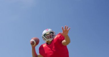 Düşük açılı, karışık ırklı bir Amerikan futbolcusunun mavi gökyüzüne futbol topu fırlatırken yavaş çekimde. Stadyumda Koşu ve Spor Eğitimi.