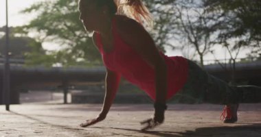 Parkta egzersiz yaparken spor kıyafetleri giymiş, yavaş çekimde şınav çeken beyaz bir kadının yan görüntüsü.