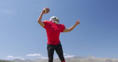 Melez bir Amerikan futbolcusunun elinde bir futbol topu tutarken, onu yere atarken ve gökyüzünü mavi bir gökyüzüne doğru ağır çekimde gösterirken ön görüntüsü. Stadyumda Koşu ve Spor Eğitimi.