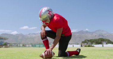 Melez bir Amerikan futbolcusunun bir spor sahasında diz çöküp futbol topunu ağır çekimde tutuşu. Stadyumda Koşu ve Spor Eğitimi.