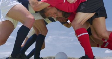 İki rakip takımdan karışık bir erkek rugby takımının düşük açılı yan görüntüsü, ağır çekimde, bir ragbi sahasında, takım forması giyiyorlar.