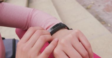 Side view mid section of a Caucasian woman on her way to fitness training, on a cloudy day, sitting, holding a yoga mat and using her smartwatch, slow motion