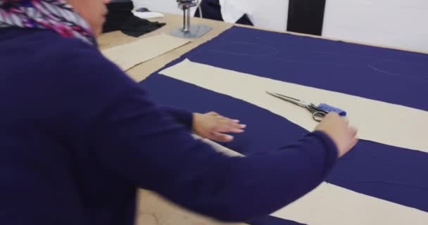 青い布の上に紙の模様が描かれた作業台に立つ混合レースの女性の肩越しに スポーツウェア工場で — ストック動画