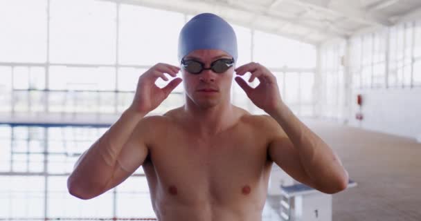 白种人男子游泳运动员戴着蓝色的泳帽 戴着泳镜 拉上护目镜 看着镜头 慢吞吞地在游泳池前观看 — 图库视频影像