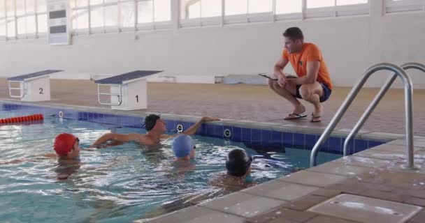 白人男子游泳教练和男子游泳运动员坐在游泳池前 沙发蹲伏在游泳池边 慢动作地与一群游泳者交谈 — 图库视频影像
